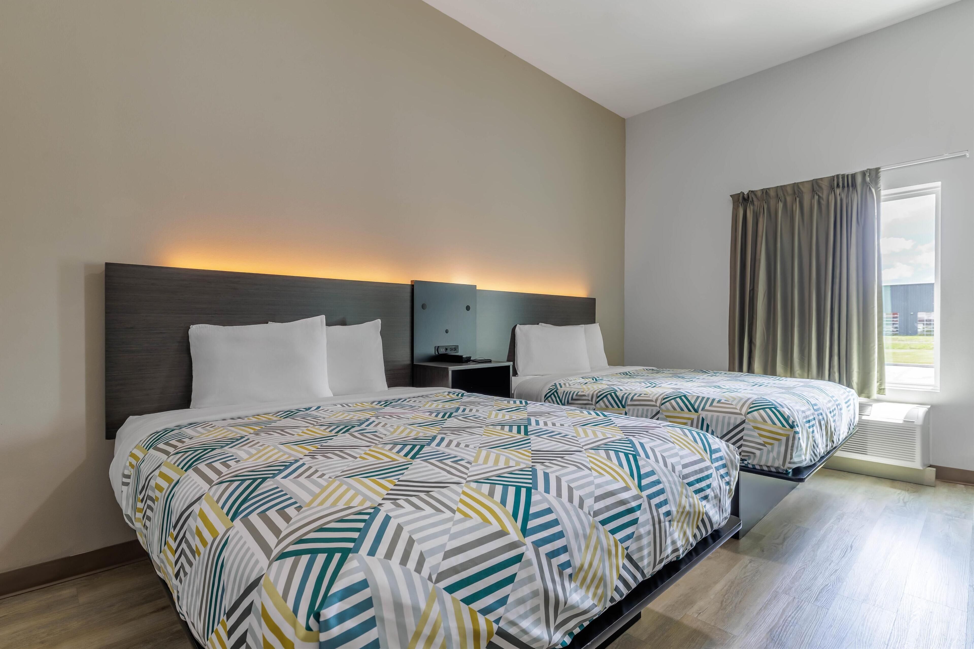 Habitació d'hotel amb dos llits, una tauleta de nit, una làmpada penjada a la paret i una finestra amb cortines.