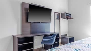 A modern hotel room with a flat-screen TV mounted on a wall, a desk with a chair, a bed with patterned bedding, and a mirror with a hanging rack.