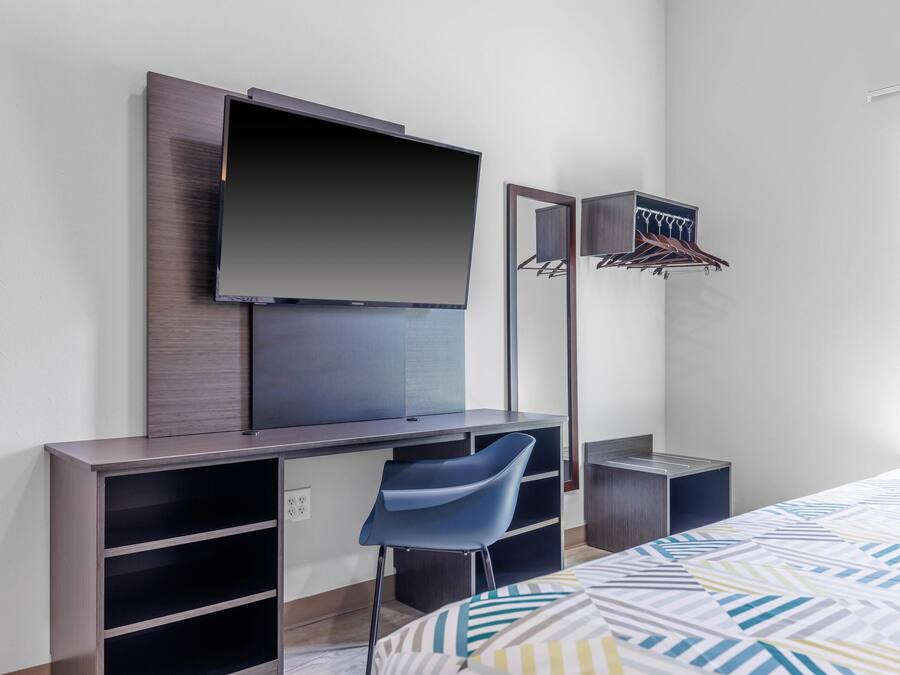 A modern hotel room with a flat-screen TV mounted on a wall, a desk with a chair, a bed with patterned bedding, and a mirror with a hanging rack.