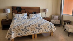 A bedroom with a large bed, two bedside tables, a chair, a small table, and a window with curtains. - Incredible views!! 1 bedroom condo with large terrace #504 at Vista Encantada! (El Divisadero)