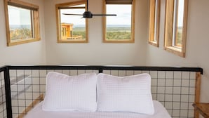 A bedroom with a bed, a ceiling fan, and three windows with a view of the landscape.