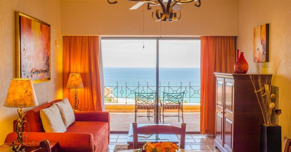 Ein Zimmer mit einem roten Sofa, zwei Lampen, einem Esstisch mit Stühlen, Meerblick und einem Balkon mit Geländer.