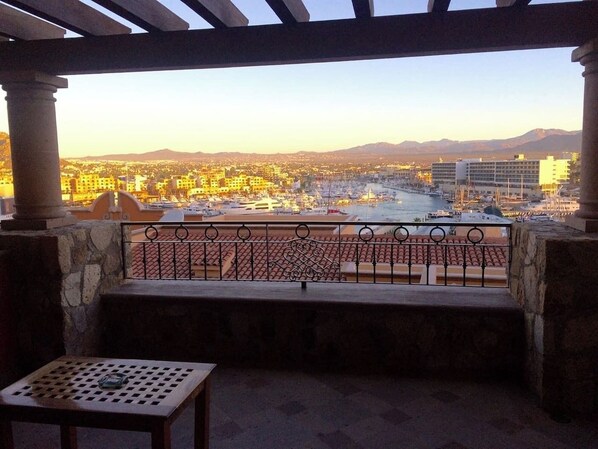 A balcony with a view of a cityscape, a stone bench, and a table with a bottle.