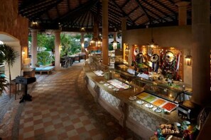 A covered outdoor area with a checkered floor, a buffet counter with various food items, and a seating area with a view of greenery.