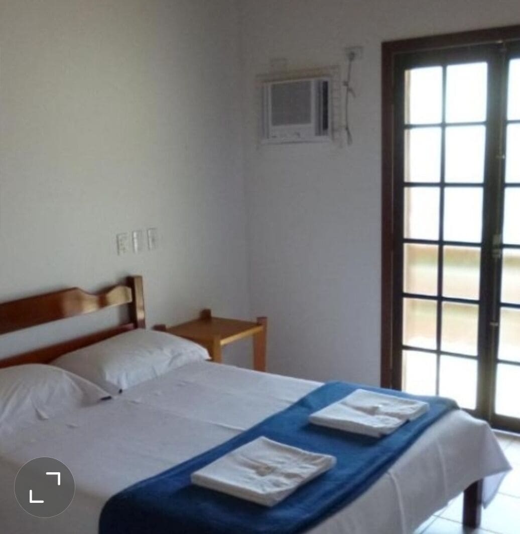 A hotel room with a double bed, a small wooden nightstand, a window with blinds, and a door leading outside.