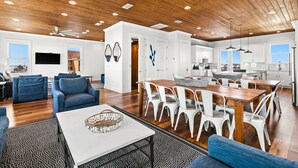 A spacious living area with a wooden ceiling, a dining table with white chairs, and a kitchen in the background.