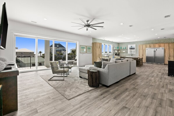 A spacious living area with a large gray sofa, a coffee table, and a dining table with chairs. There is a kitchen area with a refrigerator and a wooden cabinet. The room has a large window with a view of the outside. - Bourbon Beach - Your Dream Vacation Destination (Miramar Beach)