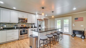Una cocina moderna con armarios blancos, electrodomésticos de acero inoxidable, una isla grande con taburetes y una bandera estadounidense en la pared.