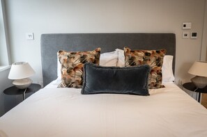 A bed with a grey headboard, white bedding, and patterned pillows. There are two bedside tables with lamps.