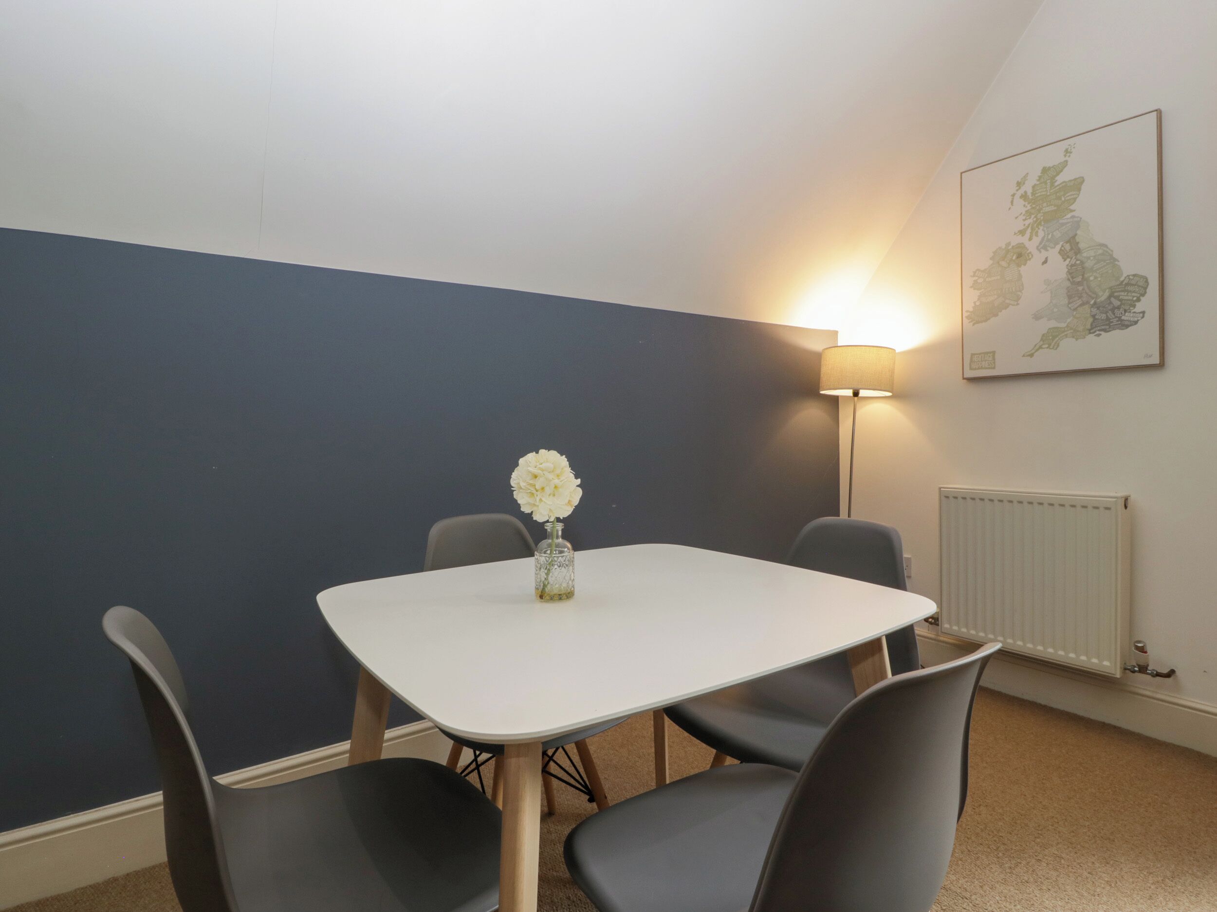 Une petite pièce moderne avec une table blanche et quatre chaises. Un vase contenant des fleurs blanches est posé sur la table. Il y a une lampe au mur et une carte du Royaume-Uni.