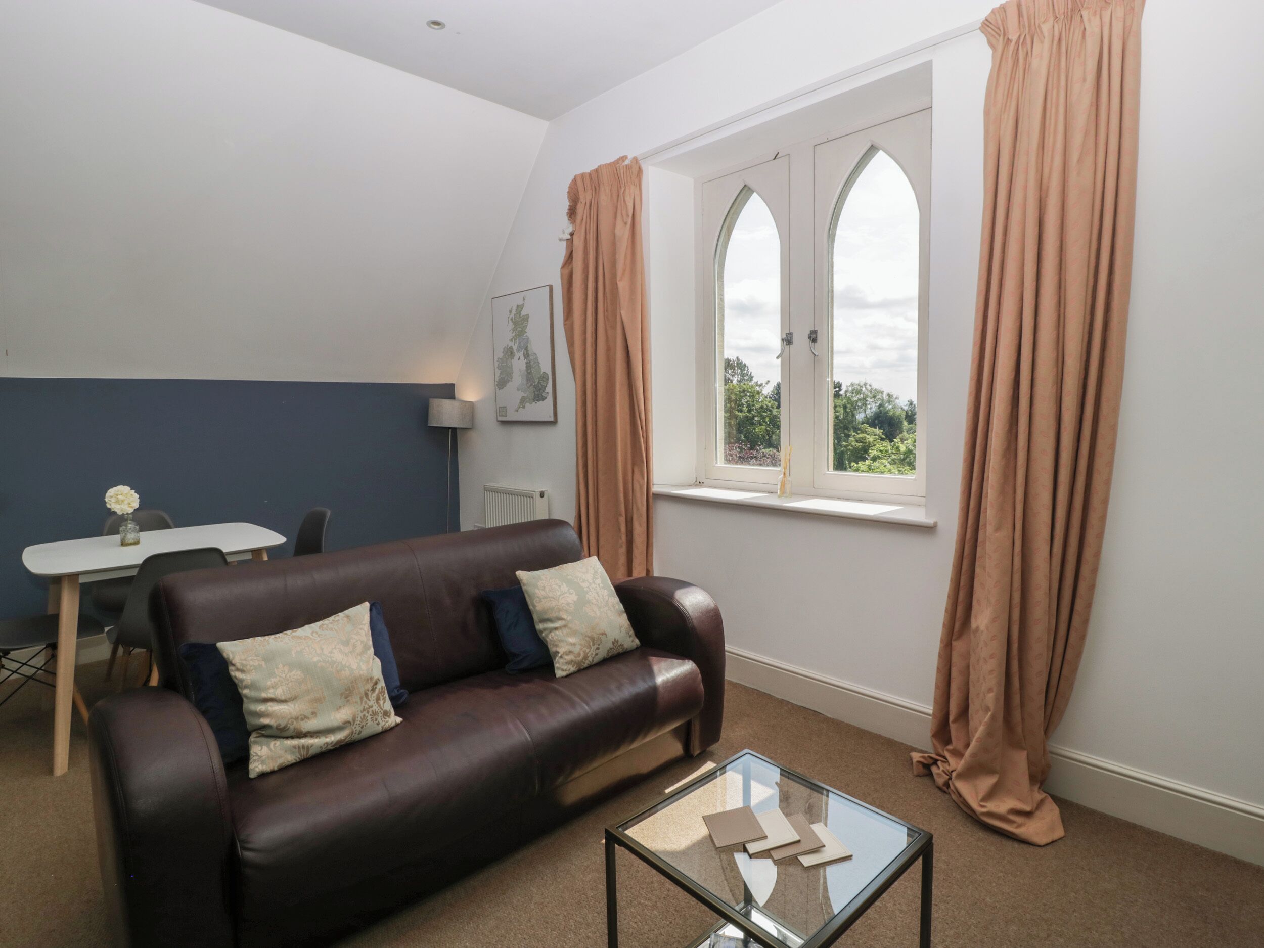 A living room with a brown leather sofa, a glass coffee table, a dining table with chairs, and a window with curtains.