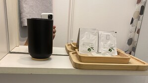 Une salle de bain avec un récipient noir, deux flacons de gel douche et de shampoing à l’extrait d’olive, et un plateau en bois.