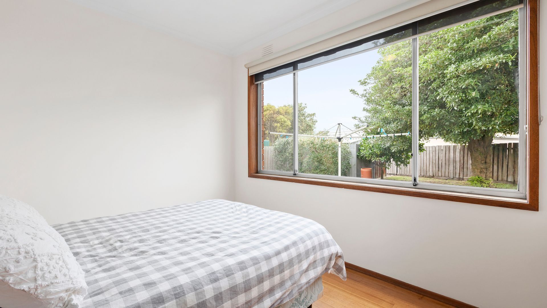 Ein Schlafzimmer mit großem Fenster, einem Bett mit kariertem Überwurf und Blick auf einen Hinterhof mit Bäumen und einer Zaun.