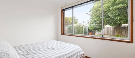 Ein Schlafzimmer mit großem Fenster, einem Bett mit kariertem Überwurf und Blick auf einen Hinterhof mit Bäumen und einer Zaun.
