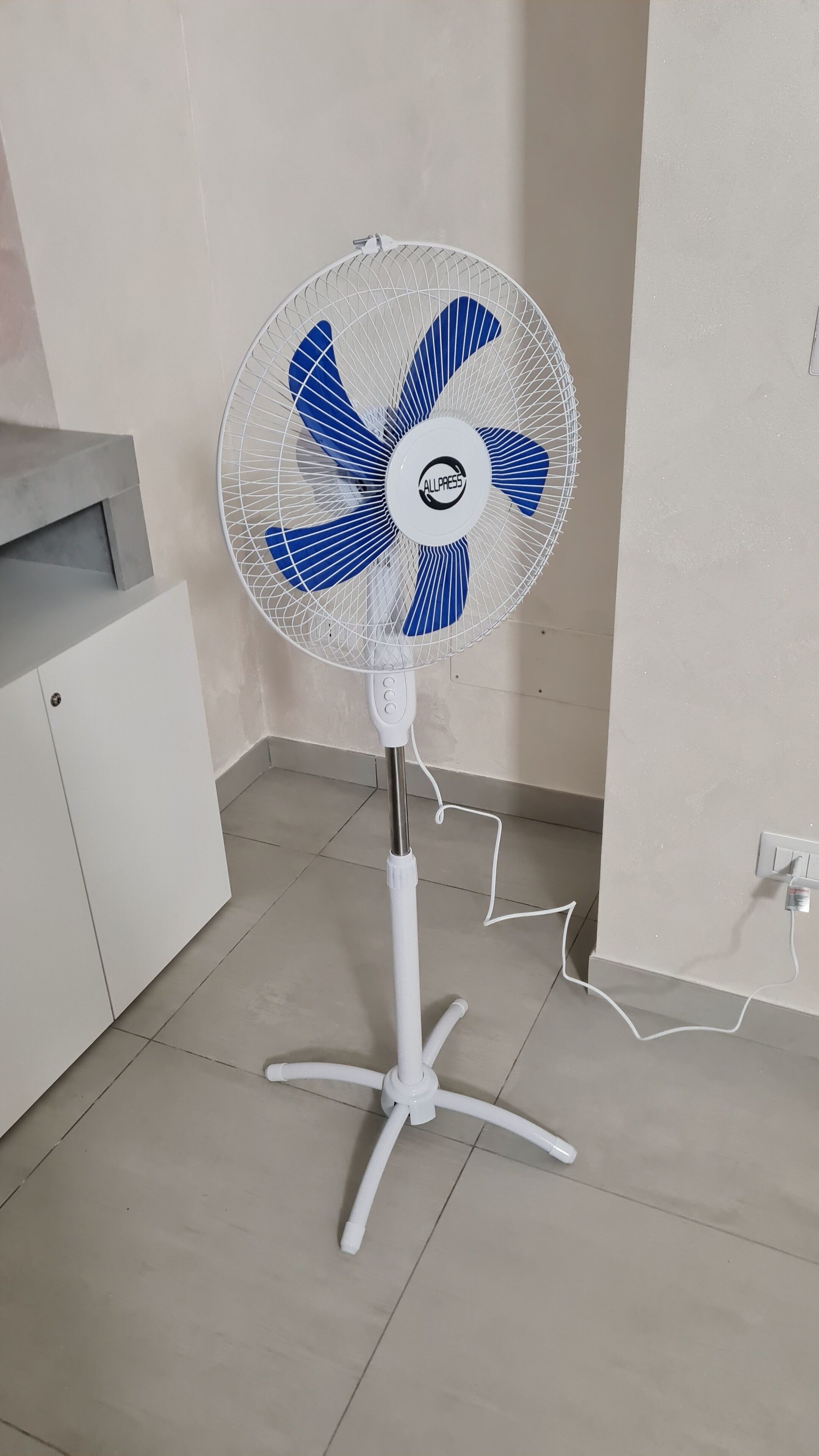 Een witte ventilator met een blauw-wit ontwerp, geplaatst op een betegelde vloer in een kamer met witte muren en een wit kastje.