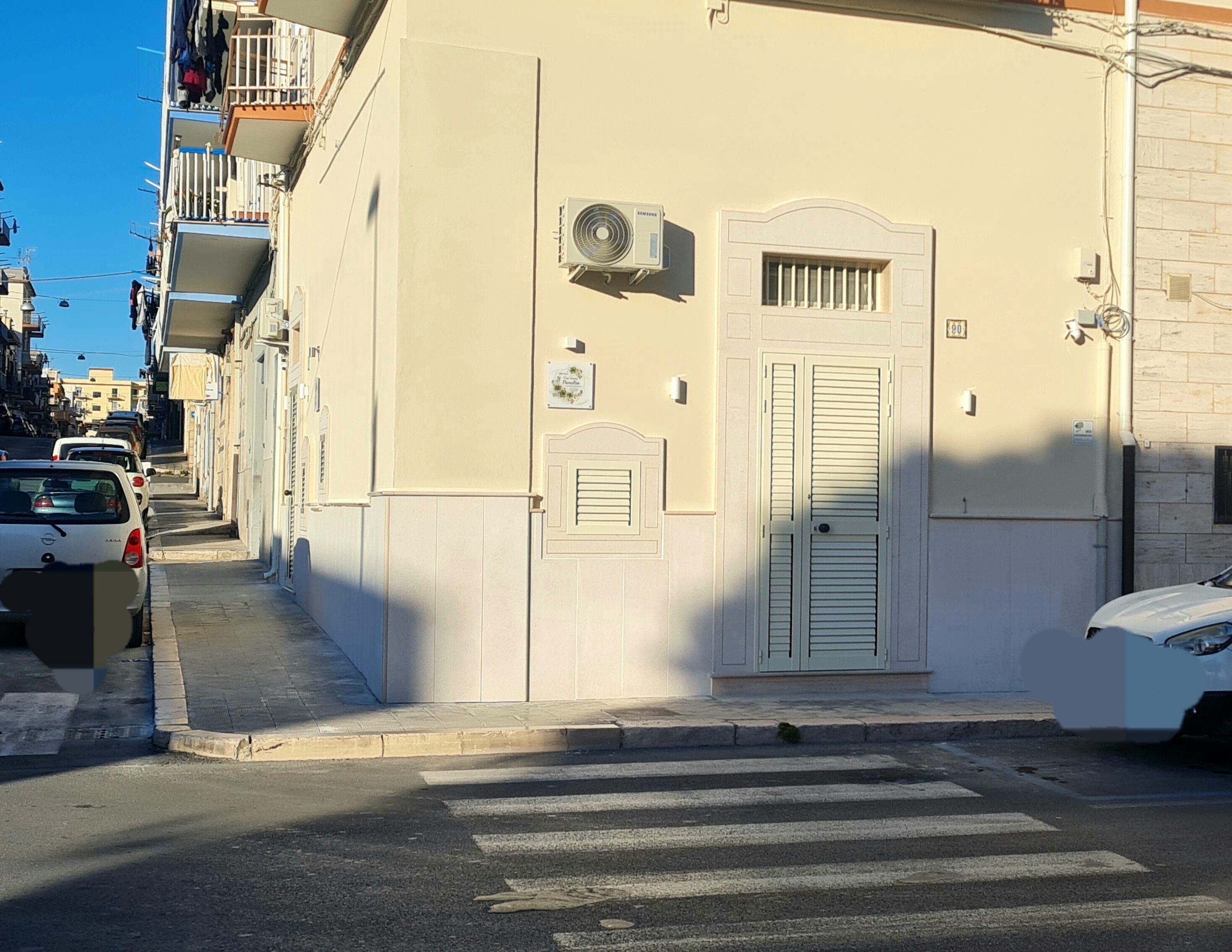 Une rue étroite bordée d’un immeuble à la façade blanche, avec une porte centrale dotée de volets et un appareil de climatisation fixé au mur.