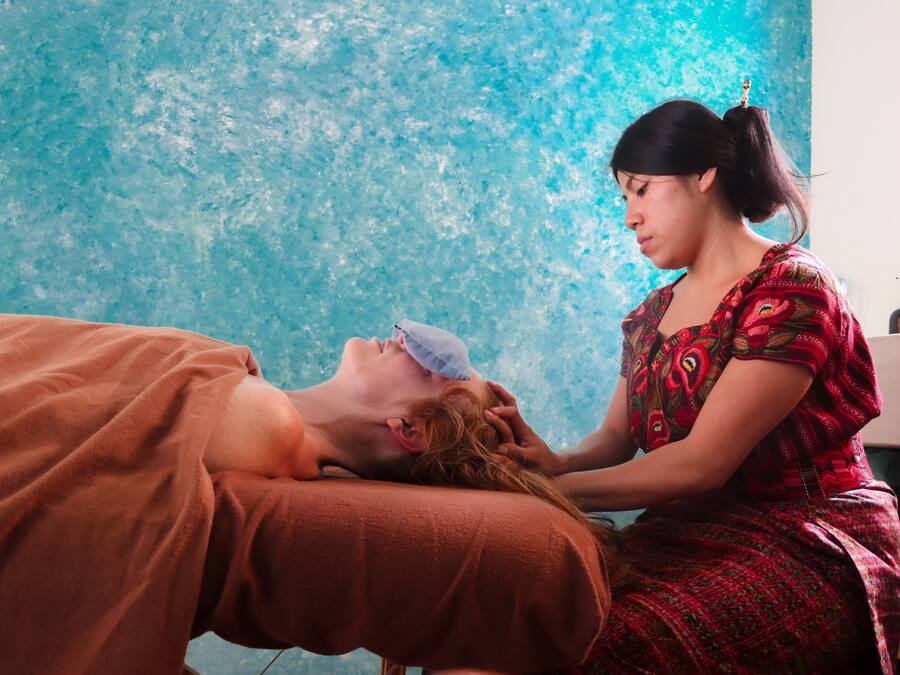 A person lying on a bed with a blanket, another person sitting beside them, and a blue textured wall in the background.