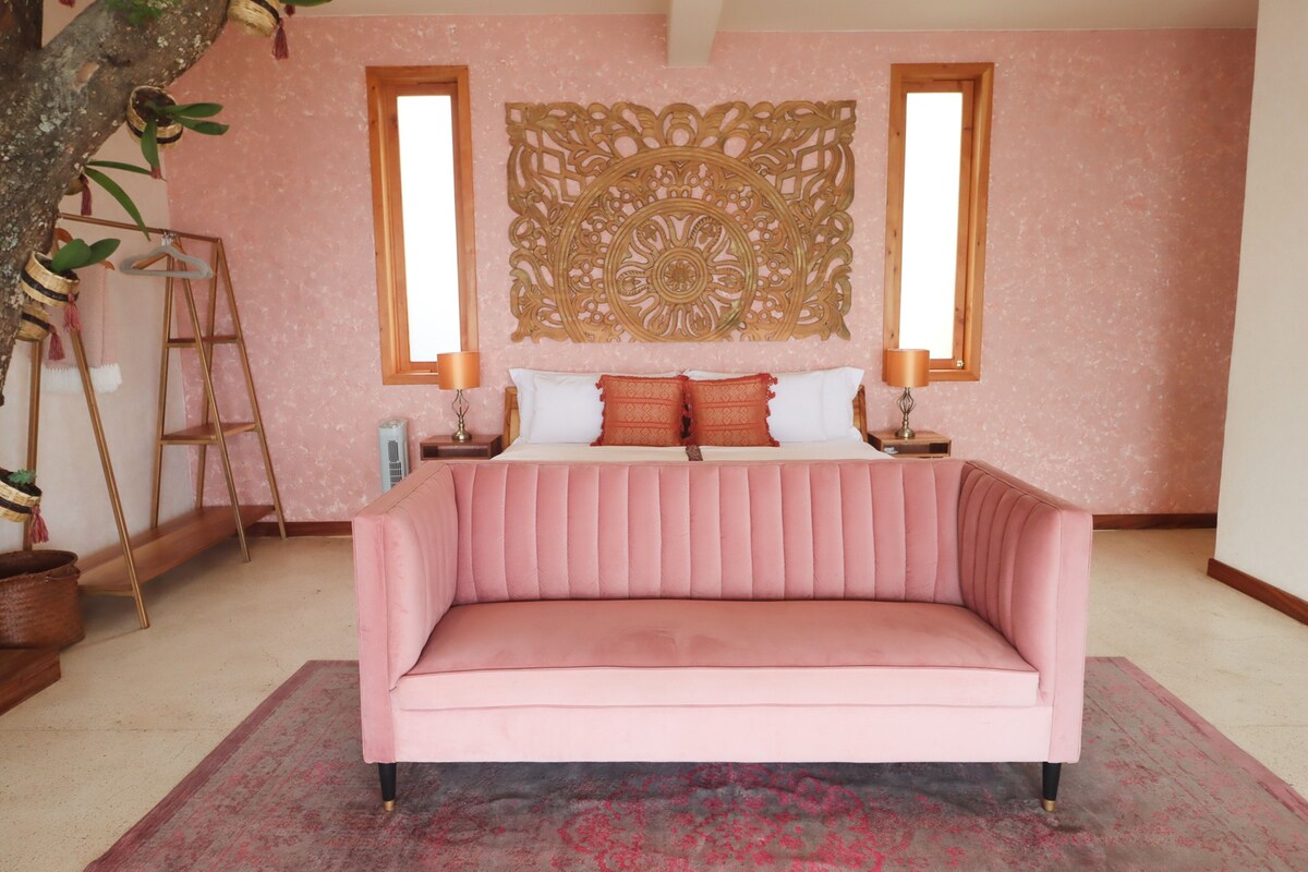 Un dormitorio con un sofá rosa, una cama con ropa de cama roja y blanca, un adorno de pared y una ventana con marco de madera.
