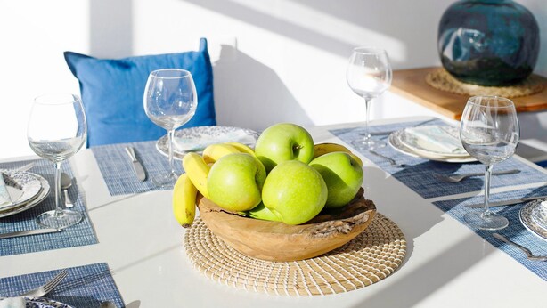 Een eettafel met een geweven placemat, een houten schaal met groene appels en bananen, en een blauw kussen op de achtergrond.
