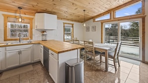 Eine Küche mit Holdekor-Schränken, einer Holz Arbeitsplatte, einem Spülbecken und einem Mülleimer. Es gibt einen Esstisch mit Stühlen und ein Fenster mit Blick ins Freie.