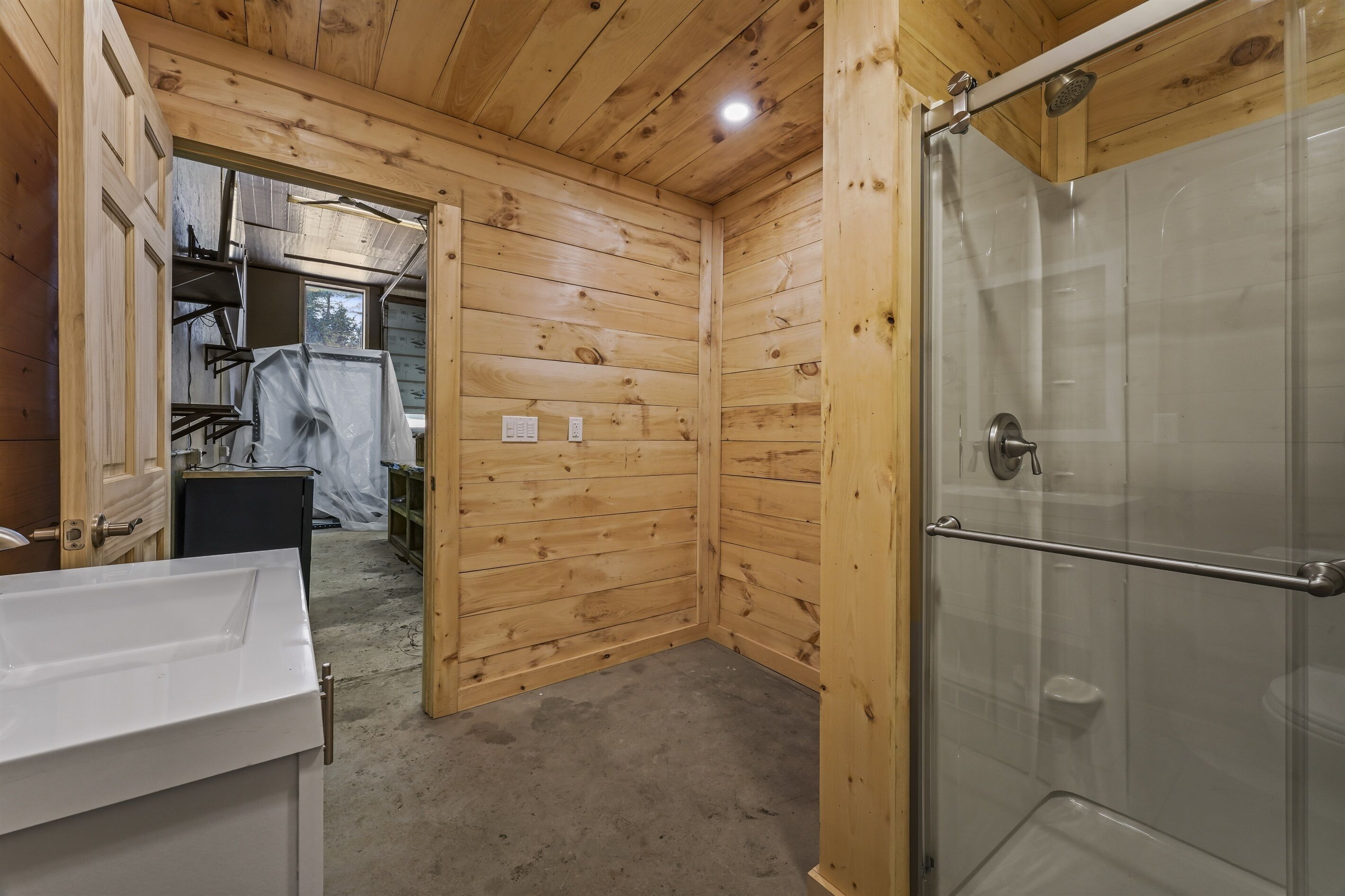 Un bagno con rivestimenti in legno, una cabina doccia in vetro, un lavandino bianco e una porta che dà su un'altra stanza.
