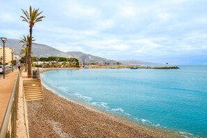 Un paseo marítimo con playa de guijarros, palmeras y vistas al mar y montañas a lo lejos.