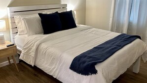 Una cama bien hecha con ropa de cama blanca y azul marino, un cabecero y una mesita de noche con una lámpara y un control remoto.