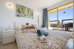 A bedroom with a bed, a bedside table, a wall-mounted artwork, and a view through a glass door.