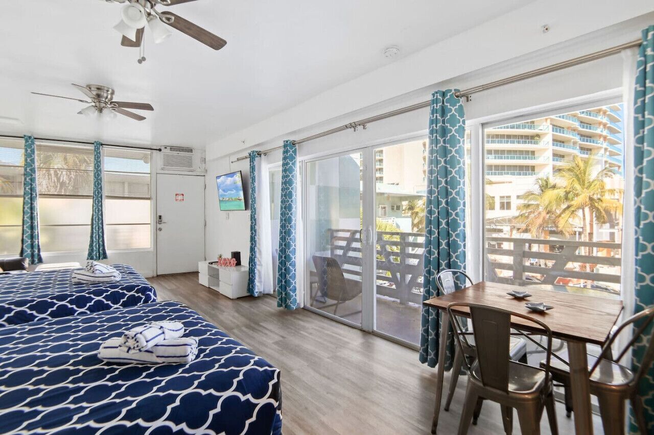 A hotel room with two beds, a dining table, chairs, a TV, and a balcony with a view of a building and palm trees.