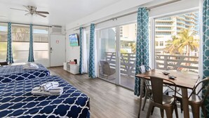 Une chambre d’hôtel avec deux lits, une table à manger, des chaises, un téléviseur et un balcon offrant une vue sur un immeuble et des palmiers.