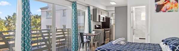A bedroom with a bed, a desk, and a chair, a kitchenette, and a balcony with a view of palm trees.