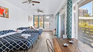 A hotel room with a balcony, two beds with patterned bedding, a wooden table, and ceiling fans.