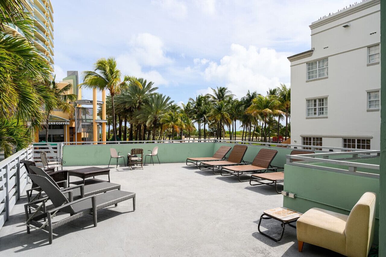 A rooftop lounge with lounge chairs and tables, surrounded by palm trees and a view of a building with a green facade.
