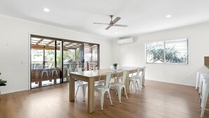 A spacious room with a wooden dining table and chairs, a ceiling fan, and a large window with blinds. - Generous Family Home with 4 Bedrooms, Pool, Pet Friendly (Sunshine Beach)