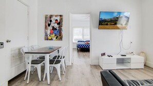Une chambre moderne avec une table à manger, des chaises blanches, un téléviseur fixé au mur et un lit recouvert d’une couvre-lit à motifs bleus et blancs.