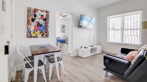 Un salon comprenant un coin repas, un canapé en cuir noir, un téléviseur fixé au mur et un grand tableau représentant Mickey et Minnie Mouse.