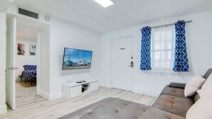 A modern living room with a grey sofa, a flat-screen TV mounted on the wall, a small white desk, and a door with a blue patterned curtain. - 2B/2B, Free Parking, South Beach, 1 Block 2 Beach (South Beach)