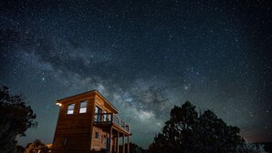 Une cabane en bois dotée d’un balcon, entourée d’arbres, sous un ciel étoilé.