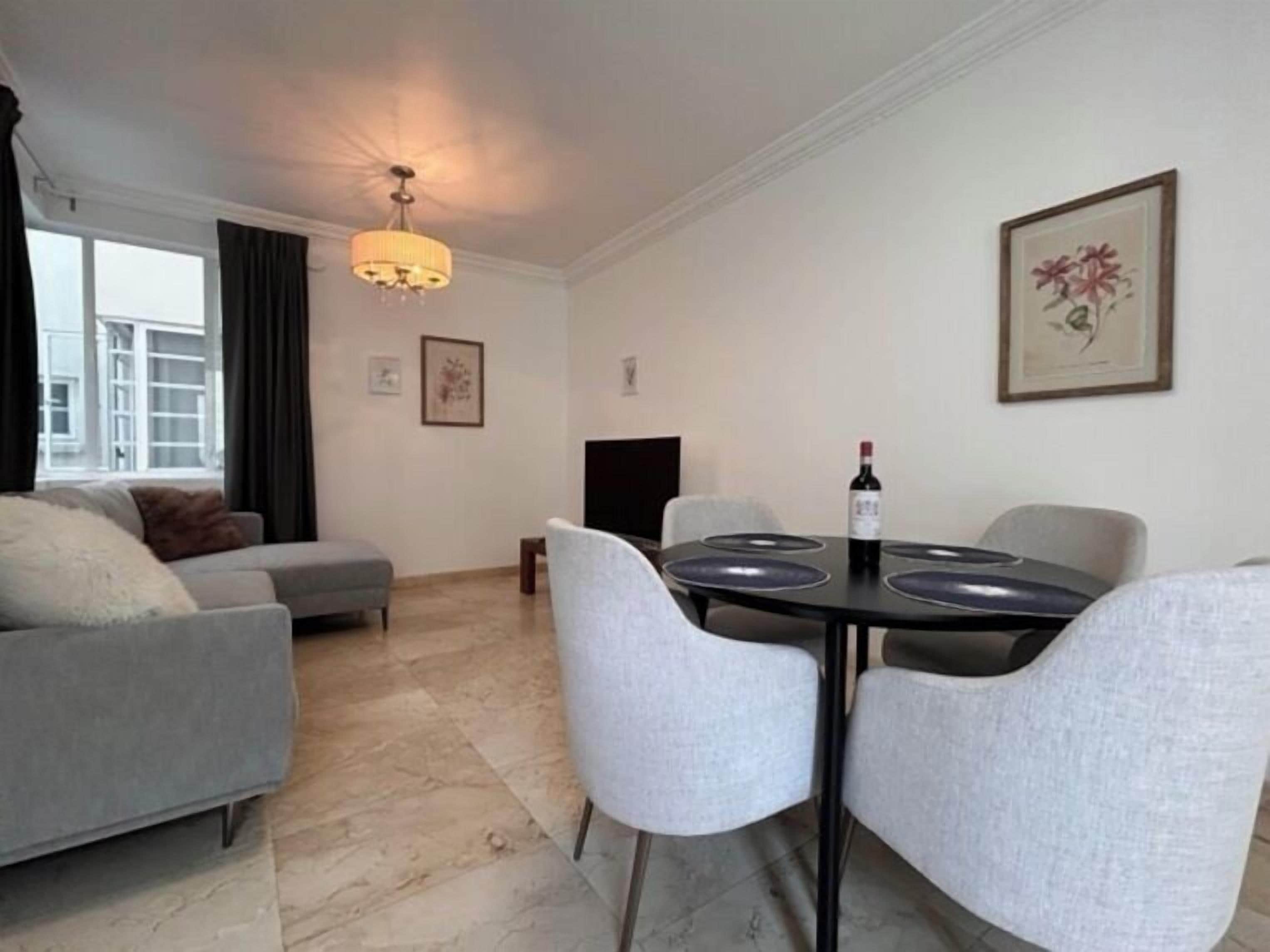 A modern living room with a grey sofa, a dining table with four chairs, a chandelier, and a framed picture on the wall.