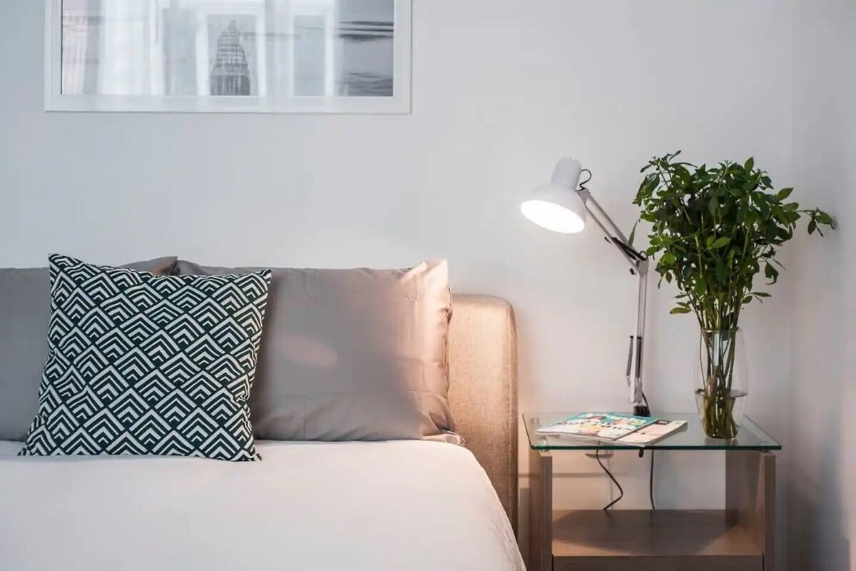 Ein ordentlich eingerichtetes Schlafzimmer mit Bett, Kissen, einem Nachttisch mit Lampe und einer Topfpflanze.
