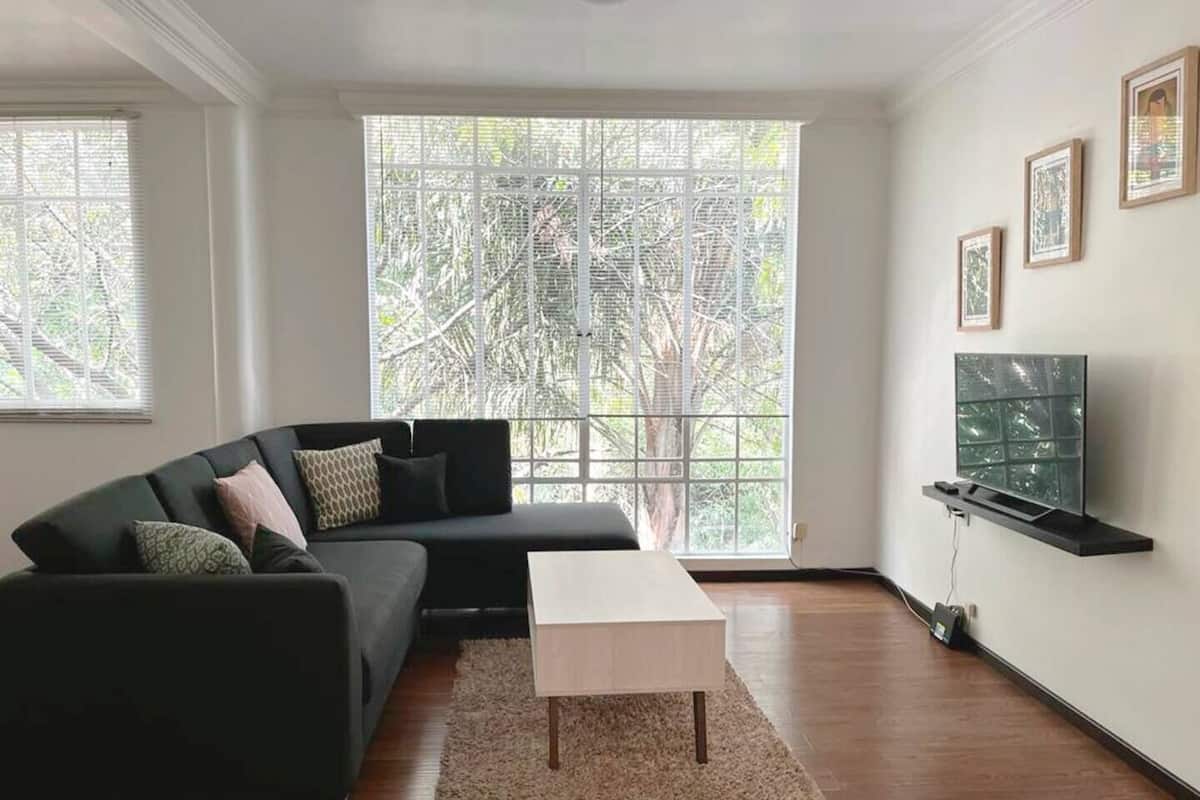 Ein modernes Wohnzimmer mit einem schwarzen Sofa, einem weißen Couchtisch und einem Fernseher auf einem schwarzen Schrank. Große Fenster bieten Blick auf Bäume.