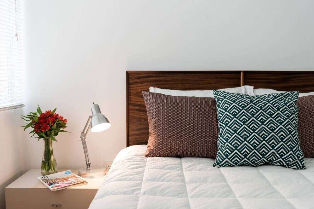 Ein ordentlich bezogenes Bett mit weißer Decke, zwei gemusterte Kissen, einer Vase mit roten Blumen und einer Nachttischlampe auf einem Nachttisch.
