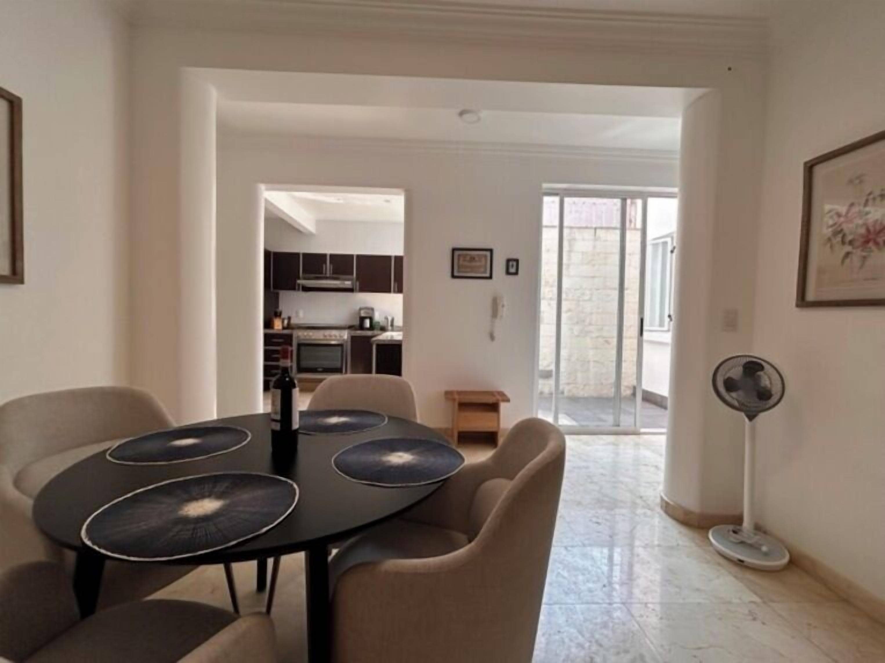 Ein moderner Essbereich mit einem schwarzen Tisch und beigen Stühlen, der in eine Küche und einen Balkon mit Ventilator führt.