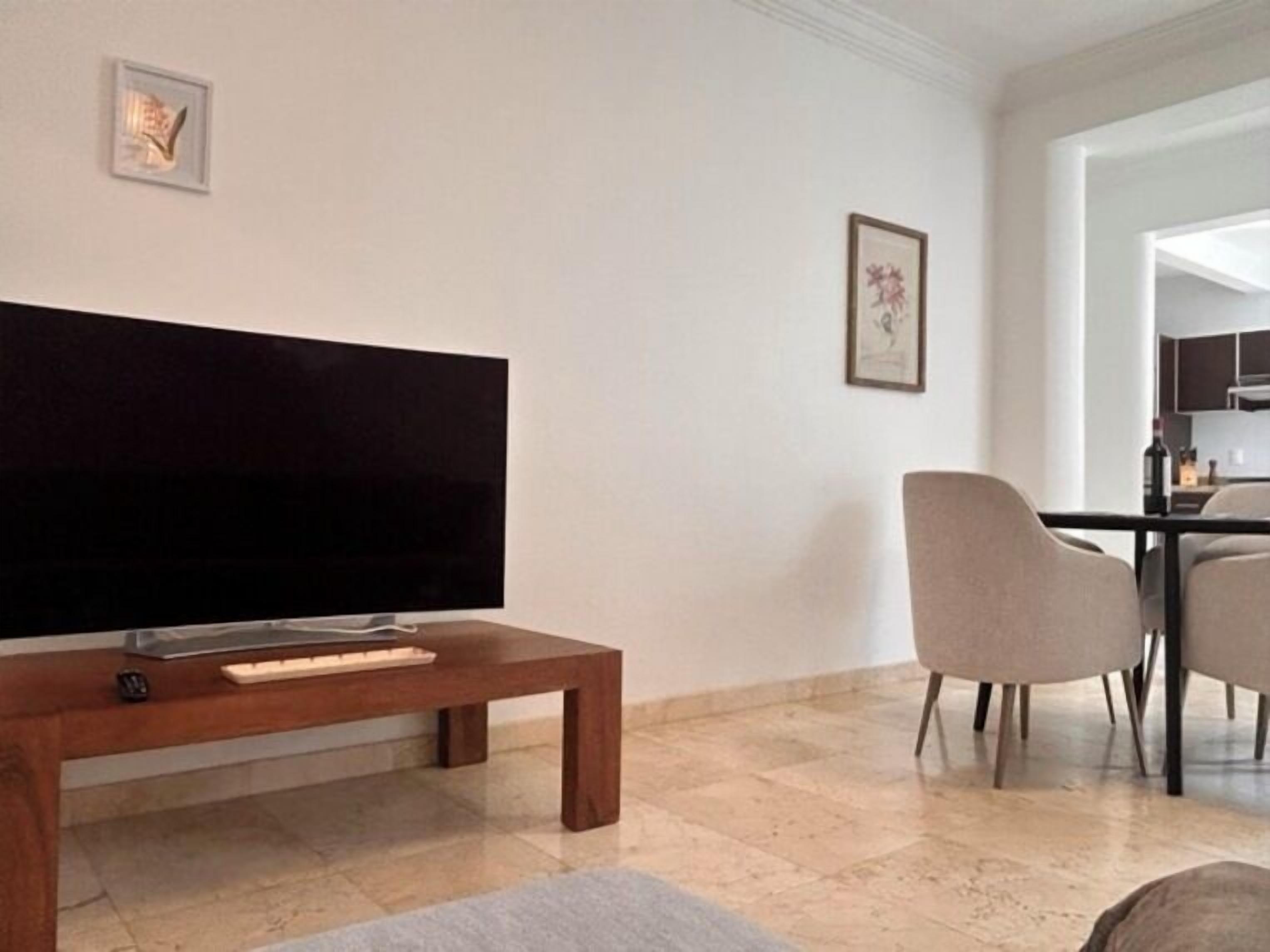 Ein Wohnzimmer mit Flachbildfernseher auf einem Holzständer, einem Esstisch mit Stühlen und einer durchgehenden Tür erkennbaren Küche.