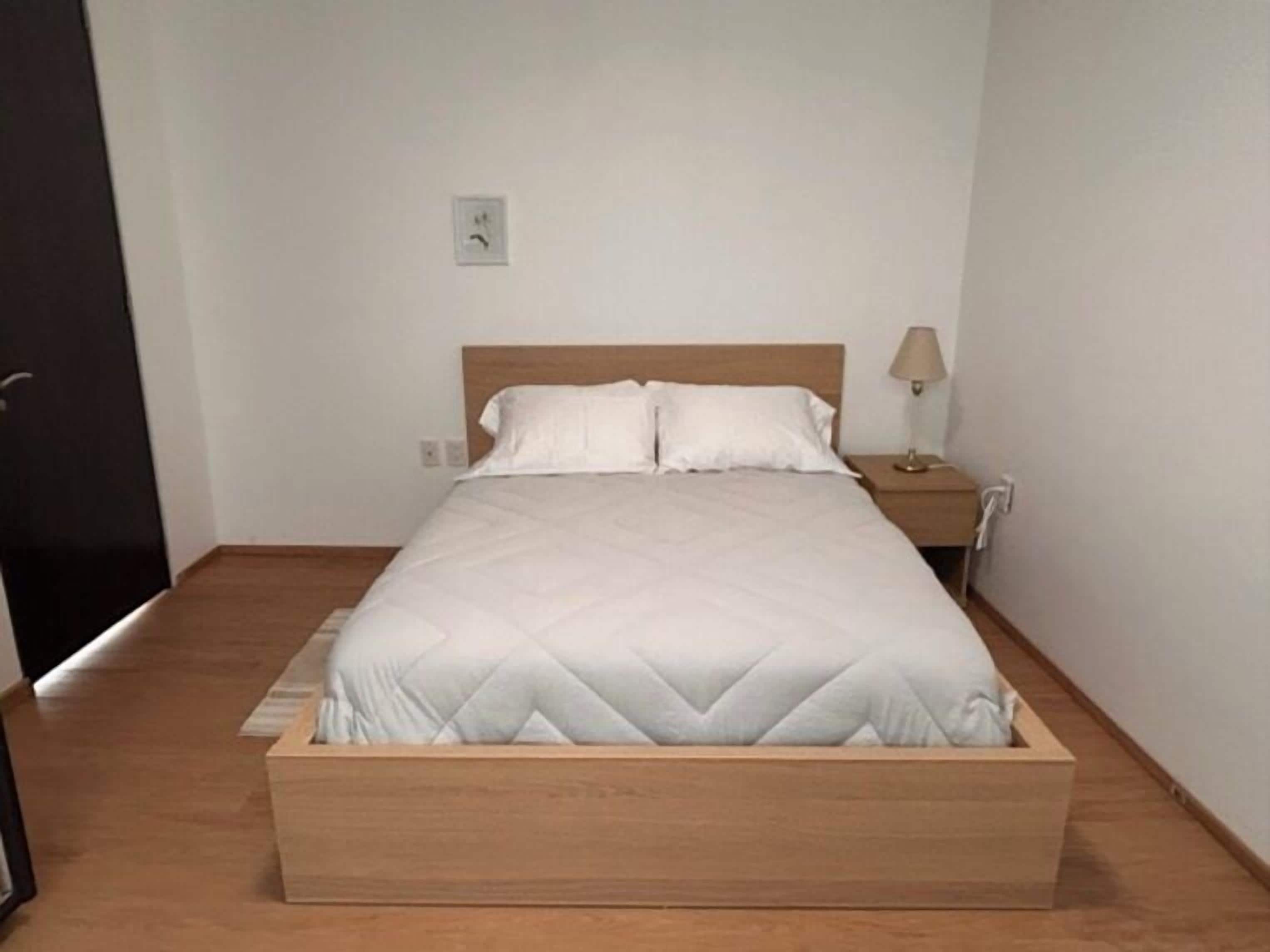 A single bed with a wooden frame and white bedding in a room with a wooden floor and a nightstand with a lamp.