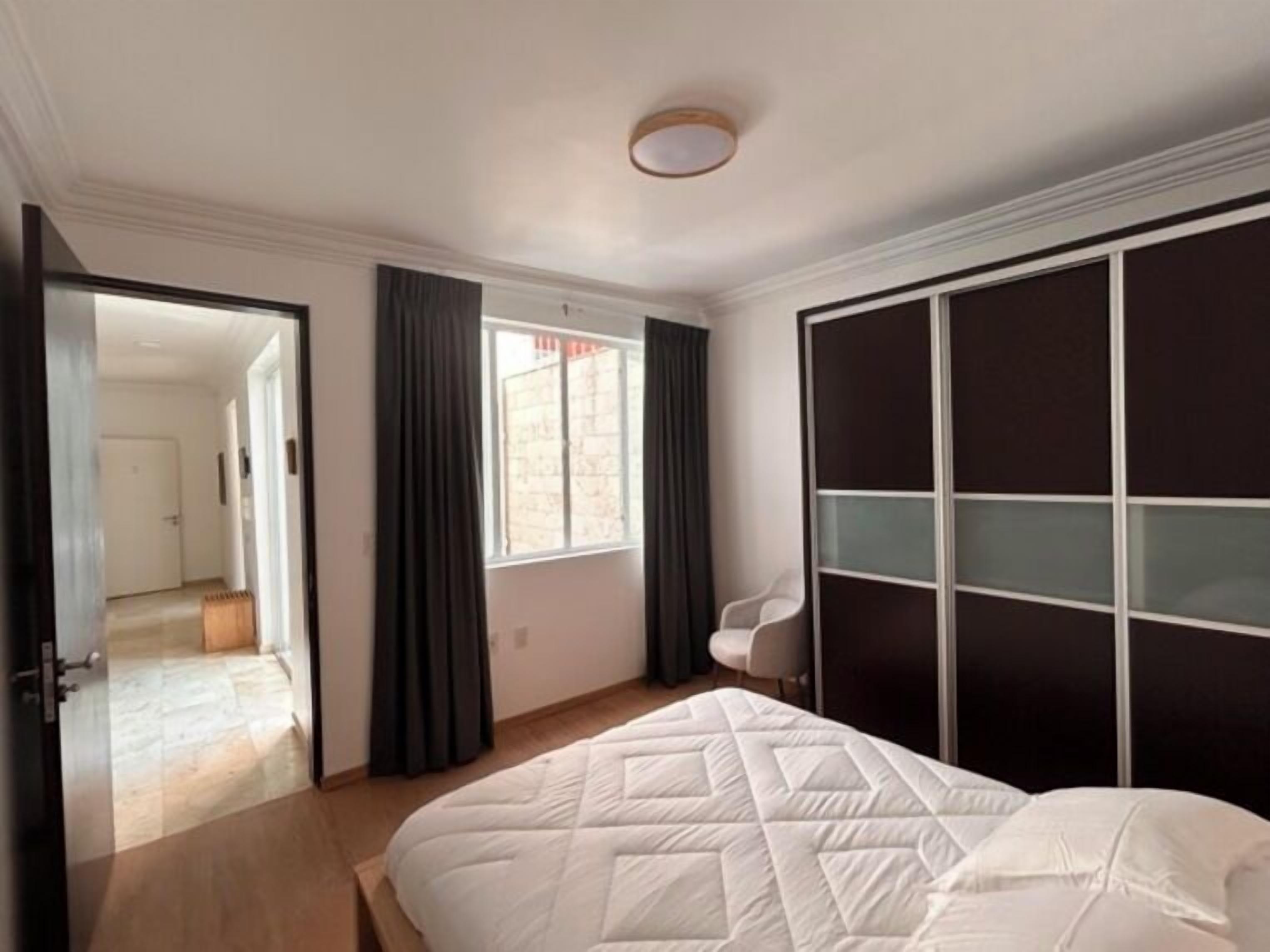 A bedroom with a bed, a wardrobe, a window with curtains, and a door leading to another room.