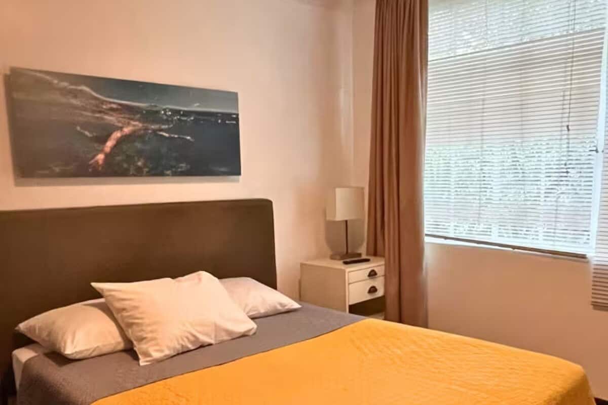 Ein Hotelzimmer mit Bett, Nachttisch, Lampe, einem Fenster mit Vorhängen und einem Bild an der Wand.