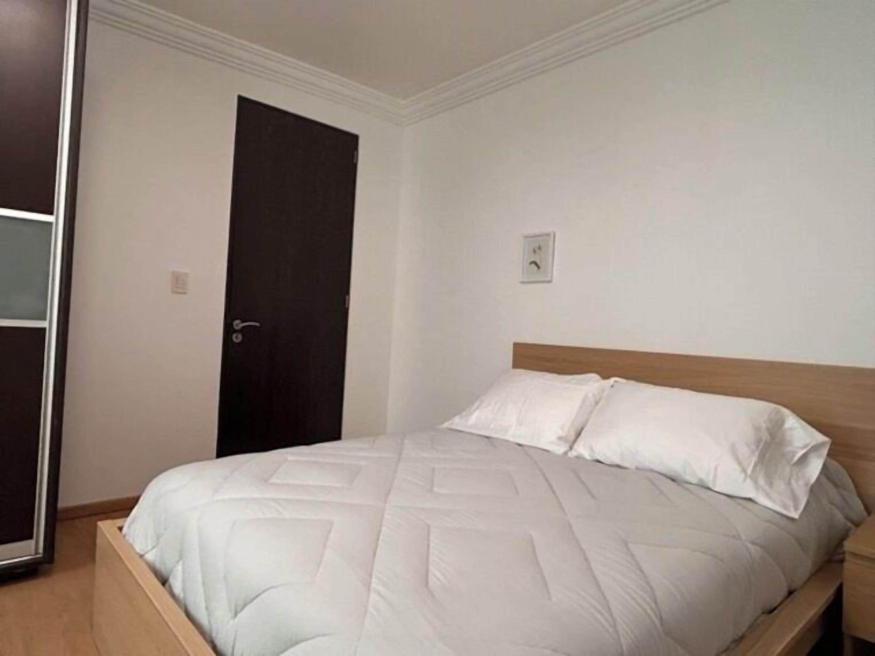 A single bed with a wooden headboard and white bedding in a room with white walls and a dark door.
