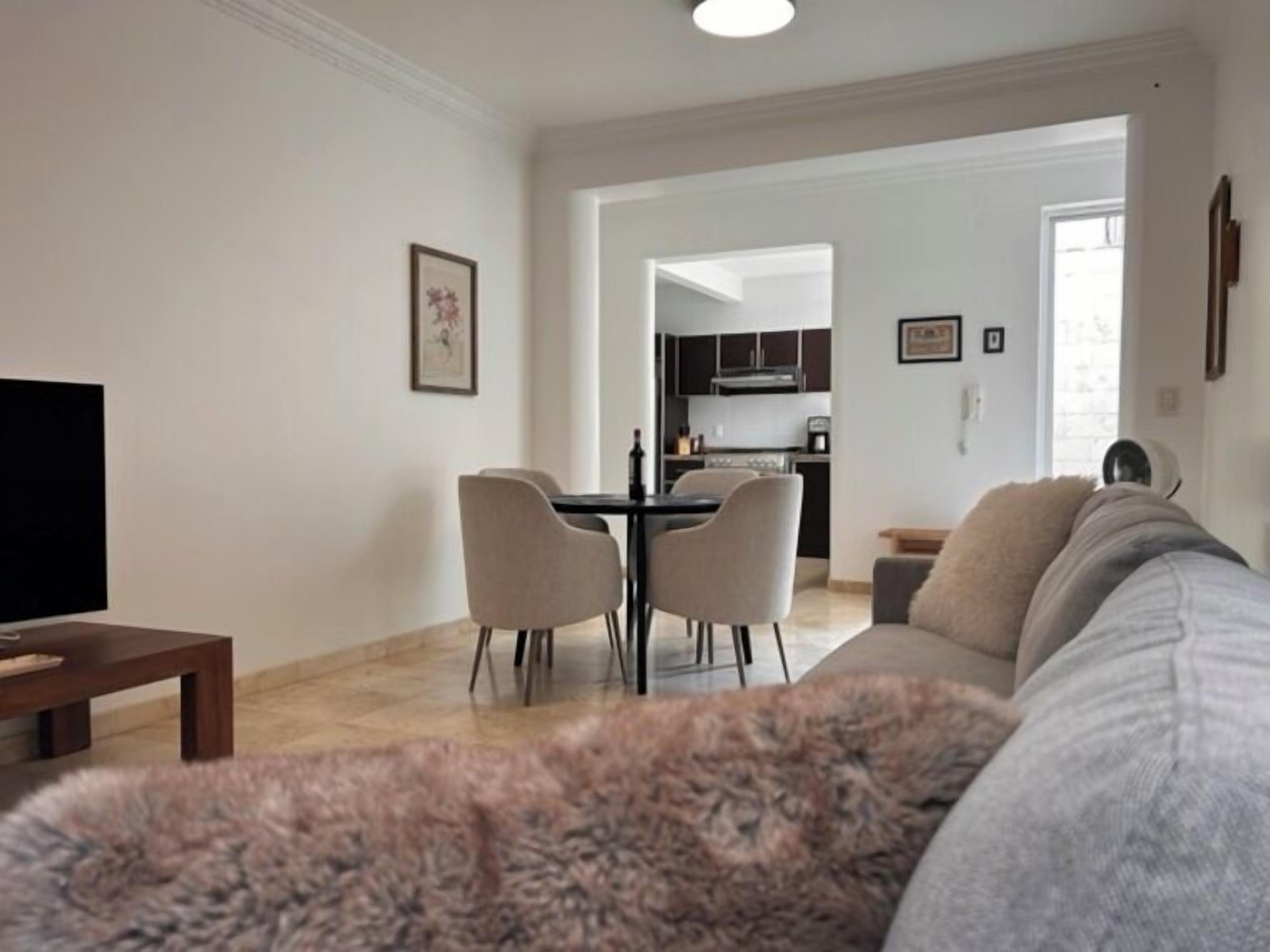 A living room with a sofa, a coffee table, and a TV. There is a dining area with a table and chairs. A kitchen is visible in the background.