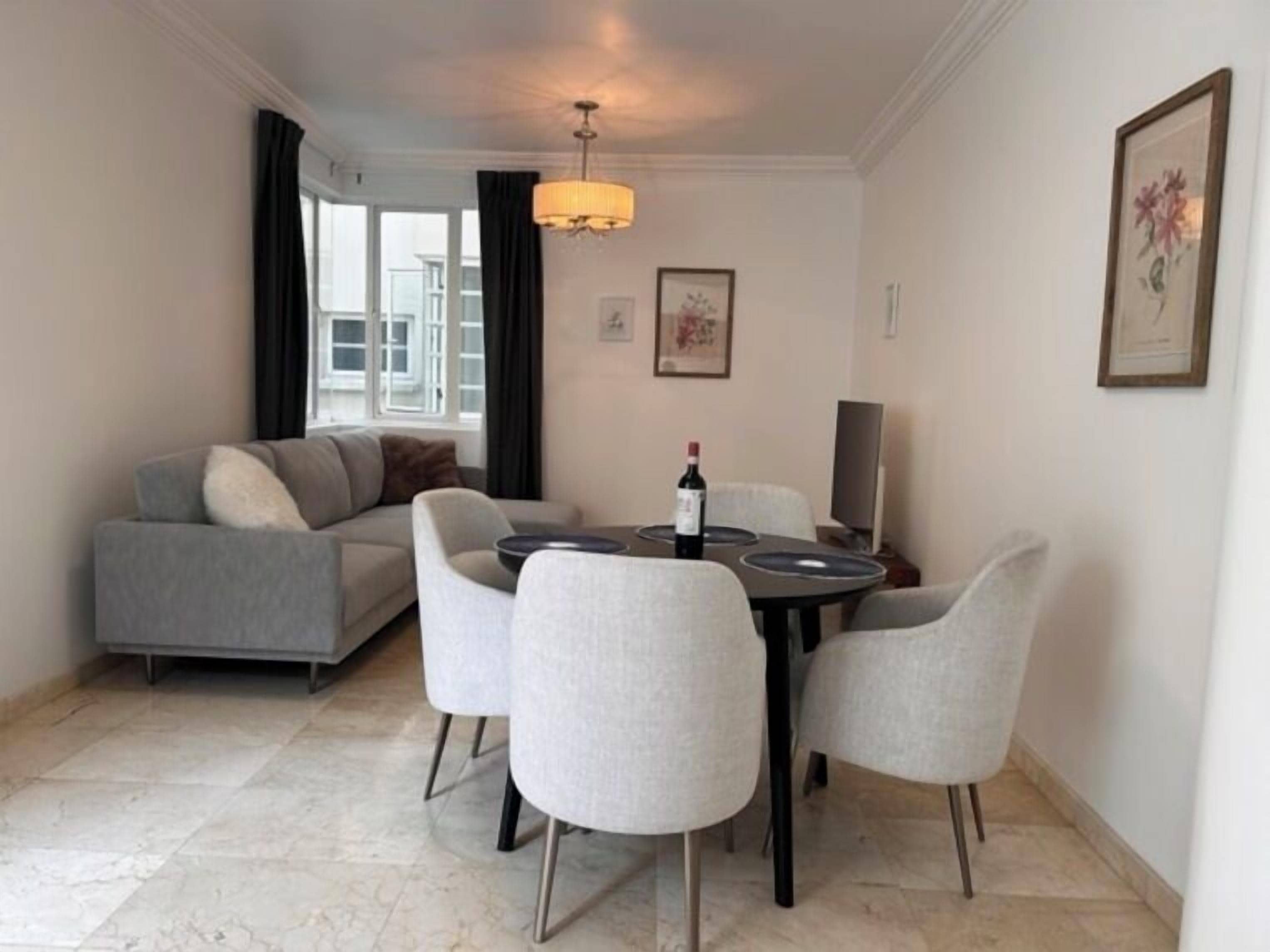 A modern living room with a grey sofa, a dining table with four chairs, a chandelier, and a framed picture on the wall.